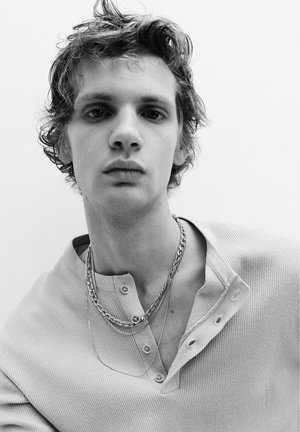 Jonge man met warrig haar, die gelaagde kettingen draagt en een geribd, dichtgeknopt shirt, kijkt recht vooruit met een neutrale blik.