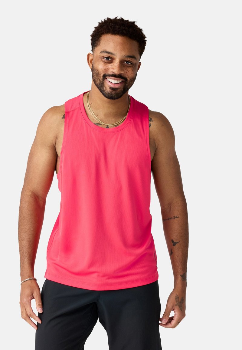 Uomo sorridente con tatuaggi che indossa una maglietta da palestra senza maniche di colore rosa acceso e pantaloncini neri, in piedi davanti a uno sfondo chiaro e uniforme.