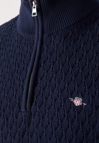 Pull en maille bleu marine avec motif en diamant, col demi-zip et petit logo écusson brodé rouge, blanc et bleu.