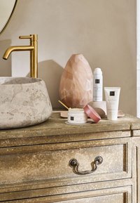 Rund, lys grå steinbassin, gull kran, rosa mønster vase, hvite hudprodukter og en teksturert gull skuffeenhet.