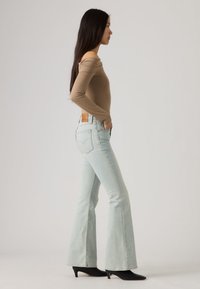 Γυναίκα σε πλάγια στάση με μπεζ τοπ έξω από τους ώμους, ανοιχτό μπλε τζιν καμπάνα και μαύρα κοντά μπότες με μυτερά τακούνια.