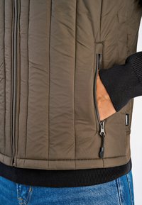 Vadderad olivgrön jacka med dragkedja fram, sidofickor och svart ribbad mudd. Texturerad tyg med vertikala sömdetaljer.