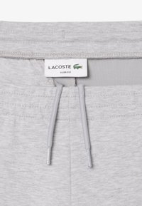 Pantalones de chándal ajustados de color gris claro con una textura suave, cinturilla elástica y cordón. Presenta un logo de Lacoste y etiqueta de marca.