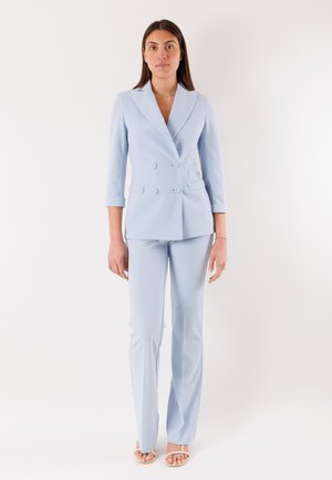 Donna in piedi, con blazer doppiopetto azzurro chiaro a maniche tre quarti, pantaloni abbinati e sandali beige con punta aperta.