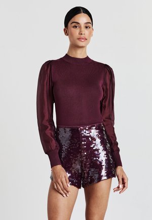 Femme portant un haut bordeaux à manches satinées bouffantes et un short à sequins assortis, se tenant devant un fond clair.