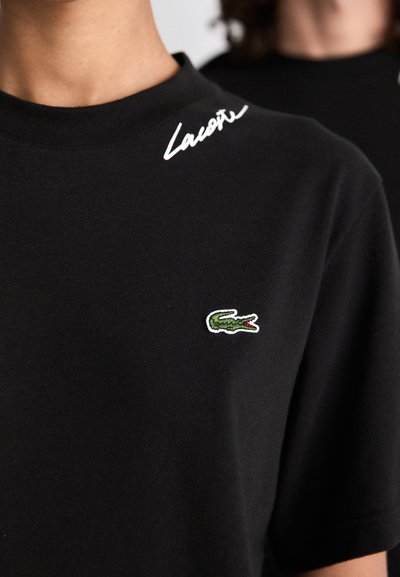 Черна тениска с бял подпис Lacoste близо до яката и малко зелено лого на крокодил на лявото гърди.