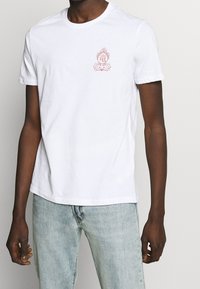 Bílá bavlněná trička s růžovou grafikou lotosu na levé straně hrudníku. Krátké rukávy a kulatý výstřih.