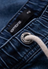 Shorts de mezclilla de color azul oscuro, con un ojal de metal visible y un cordón beige. La etiqueta de la marca dice "ombre."