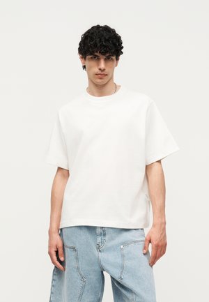 Joven con pelo rizado que lleva una camiseta blanca lisa de gran tamaño y vaqueros anchos de color azul claro, de pie frente a un fondo neutro.