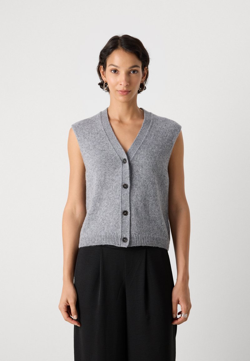 Frau in einem ärmellosen grauen, knöpfbaren Strickwesten und schwarzen, weit geschnittenen Hosen, steht vor einem schlichten, hellen Hintergrund.