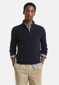 Marineblauer Pullover mit einem viertel Reißverschluss, geripptem Kragen und Bündchen; getragen über einem hellblauen gestreiften Hemd; kombiniert mit beigen Hosen.