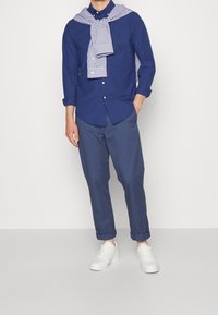 Camicia blu con bottoni sopra un maglione a righe bianche e blu, abbinata a pantaloni blu e sneakers bianche, esposta su uno sfondo neutro.