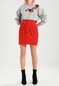 Žena s růžovými vlasy má na sobě šedou mikinu s výšivkou červené růže a červenou sukni s vysokým pasem a zavázaným opaskem, černé kotníkové boty.