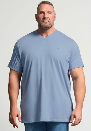 Gespiereerde man met kort haar en baard, die een lichtblauw V-hals T-shirt en spijkerbroek draagt, staand tegen een effen achtergrond.