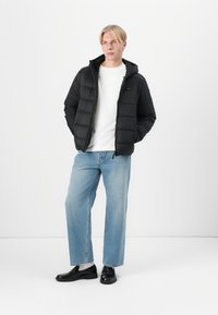 Jachetă puffer neagră cu glugă, cu design matlasat și fermoar frontal. Asortată cu blugi casual de un albastru deschis și pantofi negri.