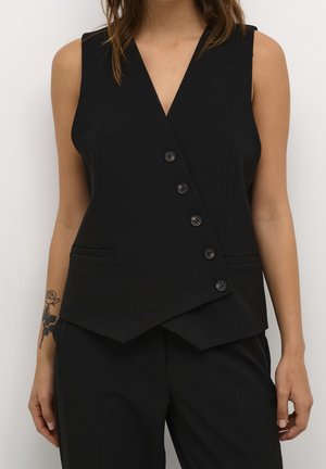 Gilet noir sans manches avec fermeture asymétrique à boutons porté sur un pantalon noir assorti sur un mannequin au bras tatoué.
