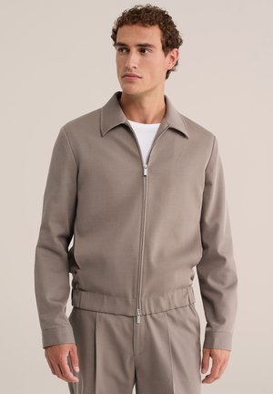 Mand med krøllet hår iført beige lynlåstrøje og matchende bukser, som kigger til siden mod en ensfarvet beige baggrund.