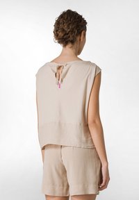 Beige, volný top s krátkými rukávy a zavazováním na zádech, vyrobený z měkké látky. Má zaoblený okraj a minimální detaily.