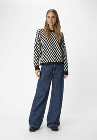 Svart och vit stickad tröja med zigzag-mönster och långa ärmar, i kombination med vida blå denimjeans och svarta skor.
