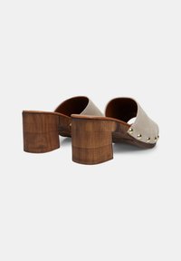 Mules en daim beige à talon bloc avec un bout arrondi et des clous dorés sur le bord extérieur. Talon en bois avec texture de grain visible.
