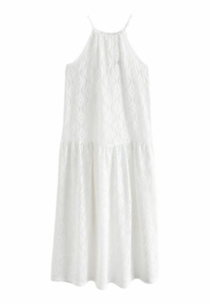 Femme portant un haut en crochet blanc à col licou avec des motifs en forme de losanges et de vagues, ainsi que de grandes boucles d'oreilles en or, sur un fond neutre.