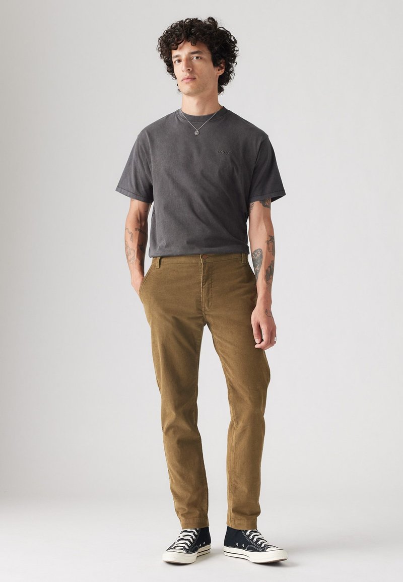Camiseta de algodón gris de manga corta, combinada con pantalones de pana marrones y zapatillas altas negras. Logotipo sutil en la camiseta.