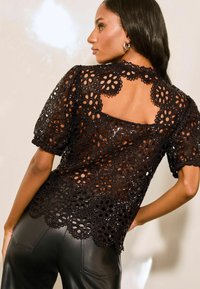 Top in pizzo nero con ritagli floreali, orlo ondulato e maniche corte a sbuffo. Indossato con pantaloni in pelle attillati.