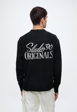 Homme portant un pull noir avec un texte blanc "Studio 90 Originals" dans le dos, associé à un pantalon blanc, dos à la caméra.