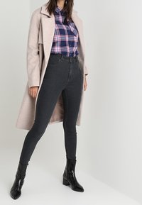 Gray skinny jeans paired with a navy blue and pink plaid shirt, layered under a light beige coat, and black heeled ankle boots.
