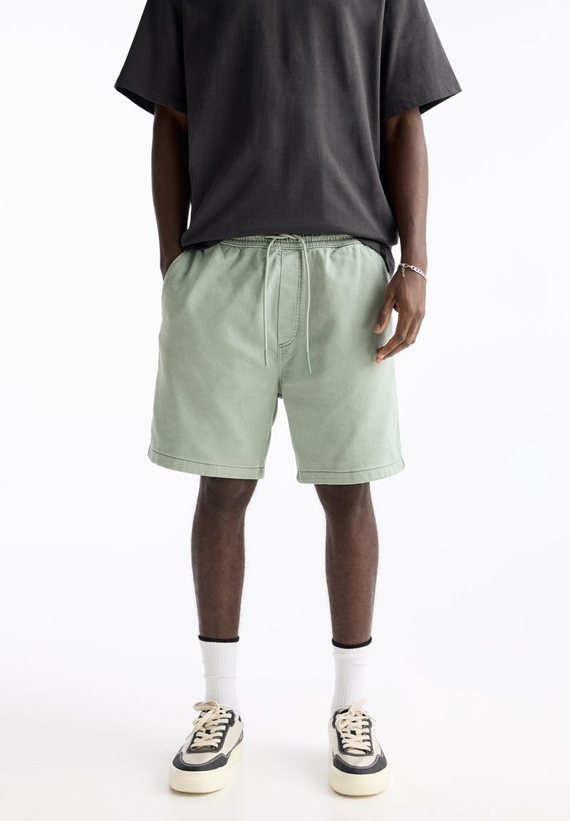 Shorts en coton vert clair avec une taille élastique et un cordon de serrage, combinés avec un t-shirt noir à manches courtes et des baskets noir/blanc.