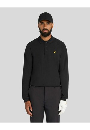 Mand med skæg iført sort kasket, sort langærmet polo med gul logo, sorte bukser og en hvid handske på højre hånd, stående foran grå baggrund.