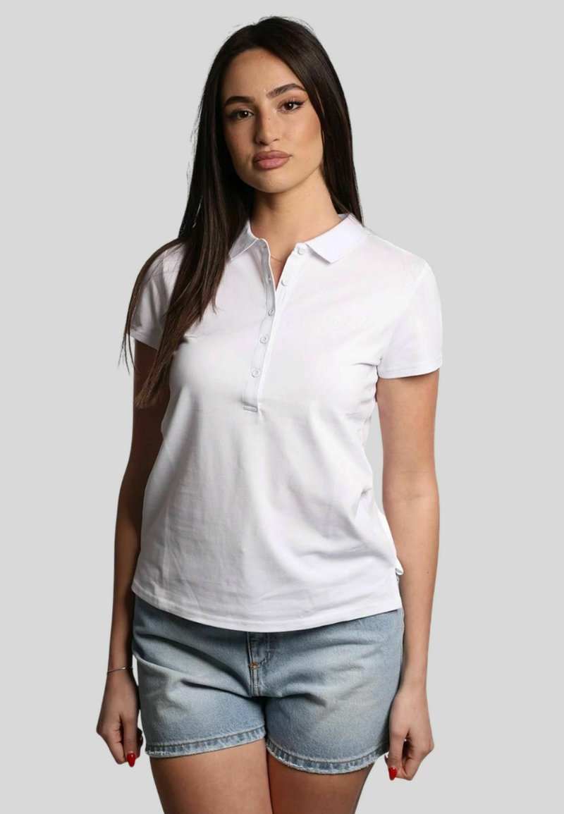 Polo bianca a maniche corte, con colletto e bottone; abbinata a pantaloni shorts in denim leggeri di colore blu. Texture liscia, vestibilità casual.