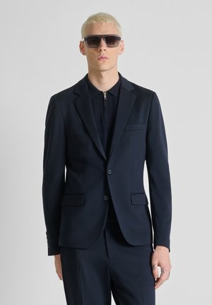 Uomo con capelli corti biondi che indossa occhiali da sole scuri, giacca da completo blu navy e pantaloni abbinati su sfondo chiaro e uniforme.