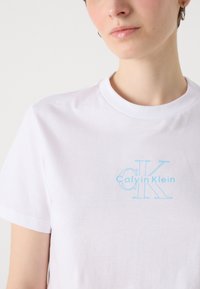 White cotton t-shirt with a crew neck, featuring a light blue "Calvin Klein" logo and "CK" graphic on the front. Smooth texture, short sleeves.