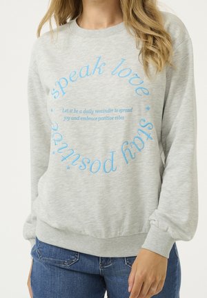 Mujer con sudadera gris claro con texto circular azul que dice "speak love, stay positive" y texto cursivo más pequeño en el interior.