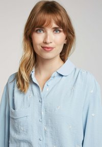 Chemise boutonnière bleu clair avec col, ornée de broderies florales blanches et d'une poche poitrine ; tissu texturé et doux.