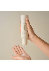 Bottiglia di shampoo cremoso, colore beige chiaro, schiacciata per erogare un liquido trasparente su una mano aperta, su uno sfondo neutro.