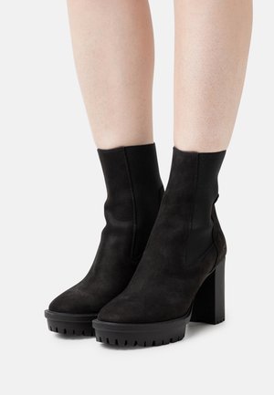 Stivaletti alla caviglia in suede nero con tacco alto spesso, pannelli elastici ai lati e suola robusta scolpita. Texture liscia e design elegante.