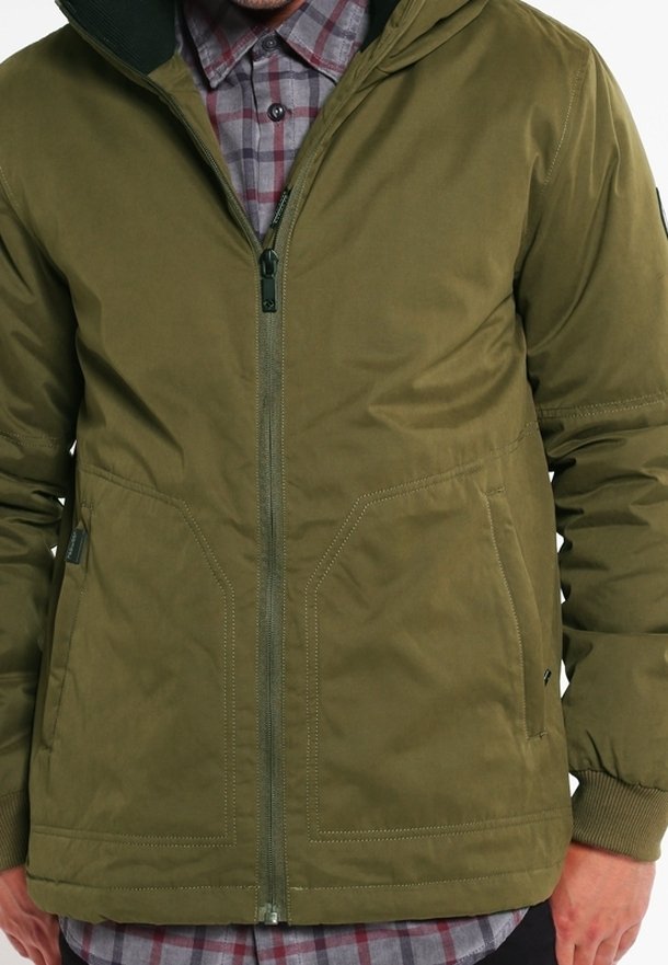 Personne portant une veste zippée vert olive par-dessus une chemise à carreaux grise et rouge avec des boutons, les mains partiellement visibles sur les côtés.