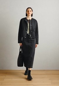 Černá zkrácená bunda s patentními knoflíky a dvěma bočními kapsami, doplněná dlouhou černou džínovou sukní. Modelka nese černou tašku a má na sobě černé boty.