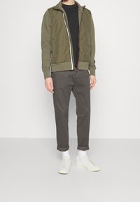 Oliwkowa kurtka bomberka z zamkiem z przodu, w połączeniu z czarnym t-shirtem. Szare spodnie chino i białe tenisówki z teksturowanymi sznurowadłami. Widoczne białe skarpetki.