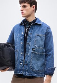 Νέος άνδρας που φοράει μπλε τζιν μπουφάν πάνω από μαύρο πουλόβερ με φερμουάρ και πουκάμισο καρό, κρατώντας μια μαύρη τσάντα στο αριστερό του χέρι.