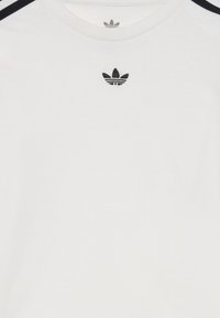Balta Adidas marškinėliai su juodu trefoil logotipu, centruotu po apykakle, ir juodomis pečių juostelėmis ant baltos medžiagos.