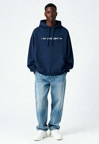 Marineblauwe hoodie met witte tekst "IK HEB JE GEZEGD DAT IK OM JE GAF," kangoeroezak, gecombineerd met lichtblauwe jeans en witte sneakers.