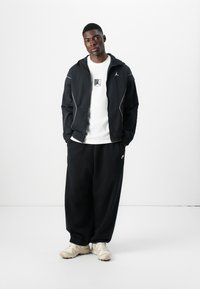 Veste coupe-vent noire avec des accents blancs, T-shirt blanc avec un logo, pantalon noir ample et chaussures de sport beige. Style simple et décontracté.