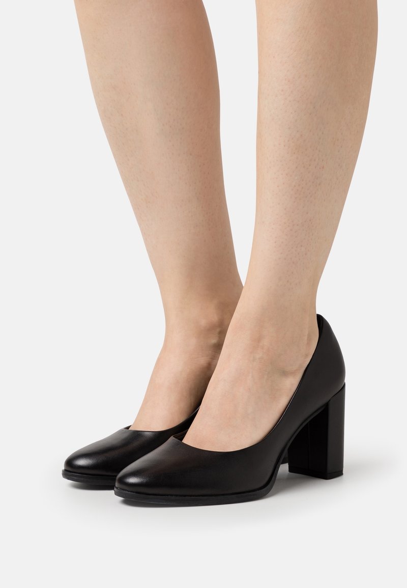 Jambes de femme portant des chaussures à bout fermé en cuir noir avec talons carrés sur fond blanc.