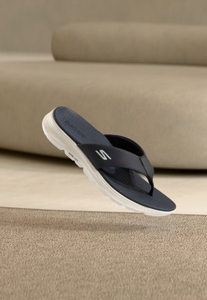 Navy blue Skechers flip-flop with white sole hovering above beige carpet, beige sofa in soft-focus background.
