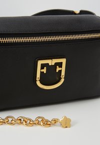 Sac en cuir noir texturé avec fermeture éclair dorée et fermoir en métal doré, à côté d'une chaîne dorée avec une petite étiquette en forme de fleur sur une surface blanche.