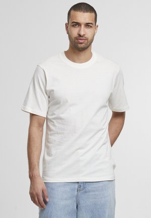 Mężczyzna z krótkimi włosami i brodą, ubrany w gładką białą koszulkę i jasnoniebieskie dżinsy, stoi z jedną ręką za plecami na szarym tle.