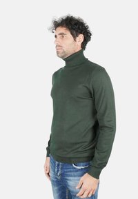 Maglione a collo alto verde scuro in morbido tessuto lavorato a maglia con dettagli a coste su collo e polsini, abbinato a jeans blu strappati.